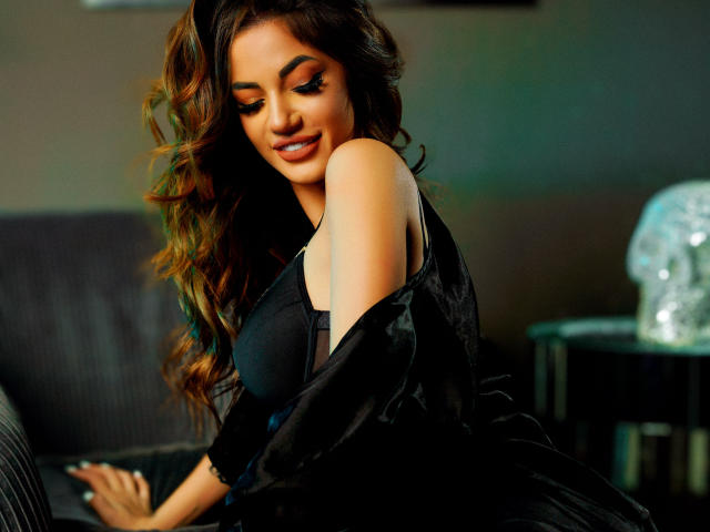 Yuukenzi with long, wavy brown hair, smiling, wearing a black satin robe, exposing her shoulder and black bra, sitting on a black couch, looking down, sexy.