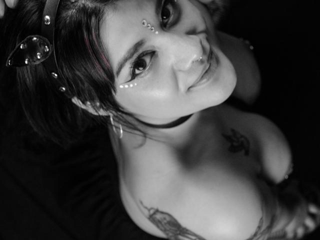 ImVioletQueen with dark hair, wearing a headband and facial decorations, looking up with a slight smile, showing large boobs and tattoos, in a black-and-white photo.