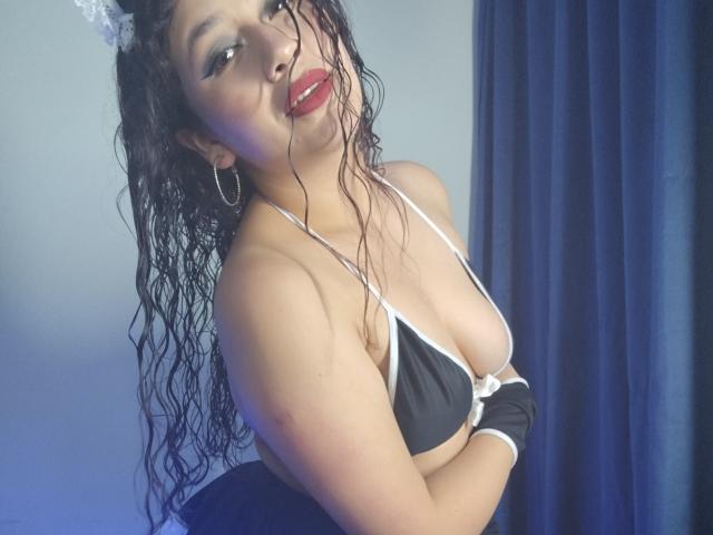 VeronicaHot69 mit nassen, schwarzen Haaren, rotem Lippenstift, einem schwarzen BH mit weißer Borte und Reifenohrringen, die ihre Brüste zeigt und verführerisch lächelt, und ein Spitzen-Katzenohr trägt..