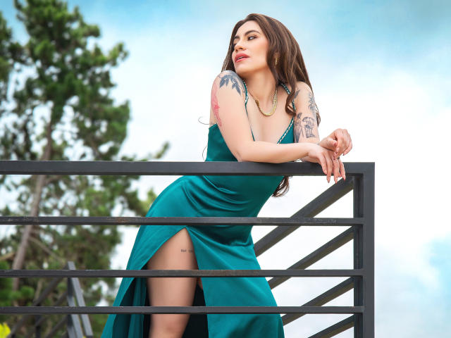 LuciaPerth in un abito verde con una spaccatura alta fino alla coscia, appoggiata a una ringhiera di metallo nero, che mostra tatuaggi sul braccio, con lunghi capelli castani e un'espressione sicura..