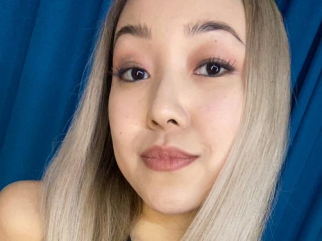RenyLime ha i capelli biondi, la pelle chiara e sta facendo una smorfia con il rossetto rosa e le lunghe ciglia. Il suo seno non è visibile. È sullo sfondo di un colore blu..