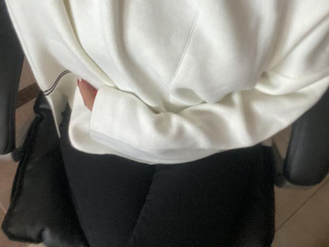 DiLarias weißer Kapuzenpullover, leicht gerüscht, bedeckt ihre Hände, während ihre schwarzen Hosen auf einem schwarzen Stuhl liegen. Ihr kleiner Finger der rechten Hand ist sichtbar..