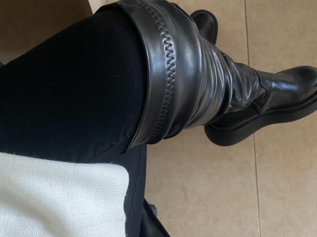 DiLarias schwarzer Lederstiefel mit aufwendigen Nähten, der am oberen Rand gerafft ist, sitzt auf ihrem schwarzen Hosenbein, über einem weißen Hemd, auf einem Fliesenboden..