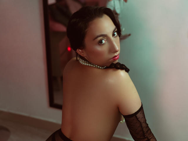 KeitlyBlue, con cabello oscuro, lápiz labial rojo, collar de perlas y guantes de encaje negro, sin sostén, mostrando su espalda y mirando por encima del hombro, exponiendo sus pechos desnudos y su trasero..