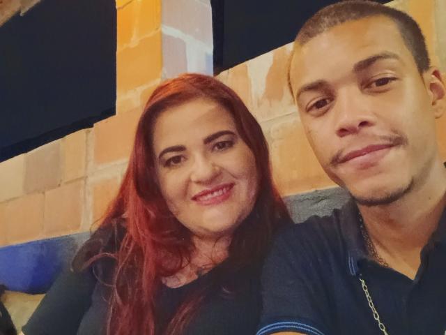 DukeDukeza, con i capelli rossi e il seno prominente, sorride, accanto a DukeDukeza con i capelli castani corti e un piccolo baffo, che indossa una camicia blu. Entrambi hanno la pelle chiara..