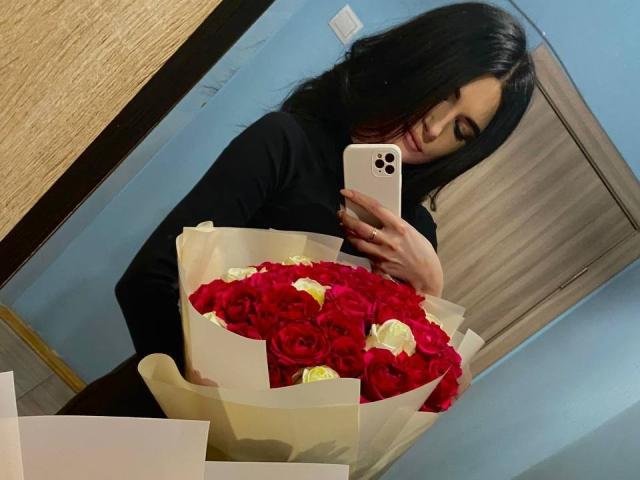 TiffanyNannette, con cabello negro, vestida con una camisa negra de manga larga, sosteniendo un ramo de rosas rojas y blancas, tomando una selfie con un iPhone..