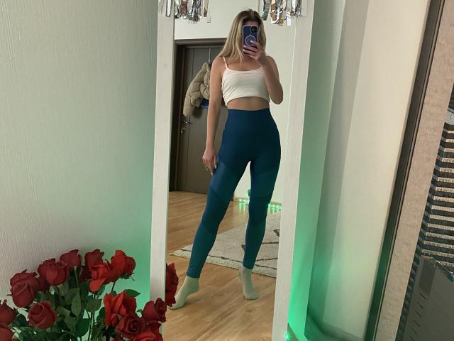 Alice, avec des cheveux blonds, un soutien-gorge de sport blanc et des leggings bleu à taille haute, tenant un téléphone, debout devant un miroir, portant des chaussettes vertes, des roses rouges au premier plan..