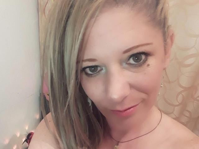SecretTemptationX ha i capelli biondi, il trucco degli occhi verde, una piccola lentiggine vicino all'occhio destro e un leggero sorriso, e indossa una collana con un piccolo ciondolo..