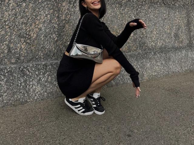 MarieAngel, hockend, trägt einen schwarzen Pullover, einen Minirock, eine metallische Handtasche, schwarze und weiße Turnschuhe, lächelt, schwarze Handschuhe ohne Finger, gebräunte Haut, kurze schwarze Haare, sichtiges Gesäß, Hintergrund: eine Betonwand..