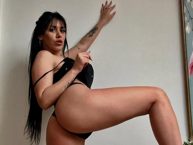LaylaCruz, avec de longs cheveux noirs, portant un string noir, levant une jambe, montrant ses fesses, avec des tatouages sur le bras, regardant l'appareil photo, devant un mur blanc..
