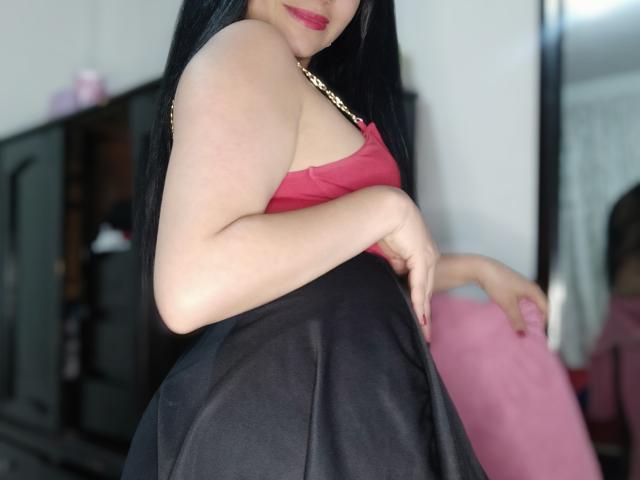SkyblueKate mit langen schwarzen Haaren, einem roten Tanktop, einem schwarzen Rock, die ein pinkes Kissen hält, ihr Hintern zeigt, einer goldenen Halskette, rotem Lippenstift und einem leichten Lächeln in einem Schlafzimmer..