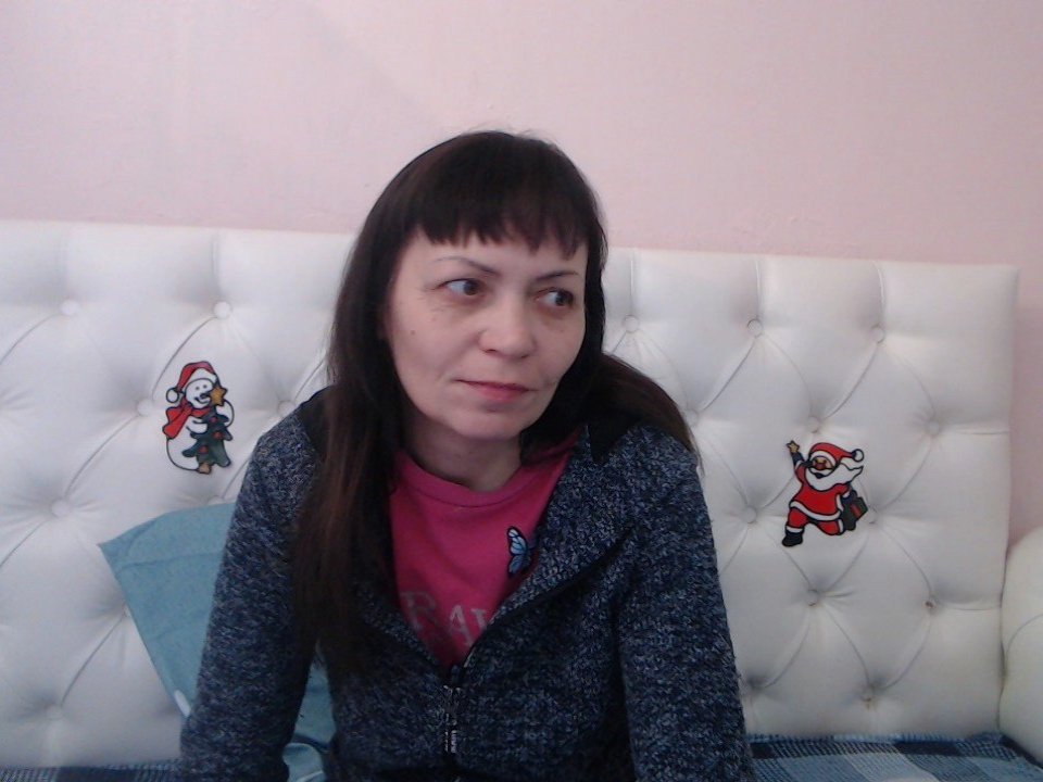 StellaHaart with brown hair, wearing a blue zip-up hoodie over a pink shirt, sitting on a white tufted bed with cartoon monkey stickers on the headboard.