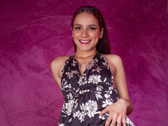 MaiaPark with brown hair, smiling, wearing a black floral halter dress, pointing with her right hand, pink lipstick, medium tits, against a purple textured wall.