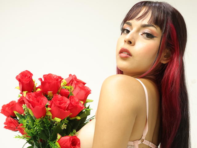 AlySnow with red roses, holding them in front of her bare boobs, has black and red hair, pink lingerie, and sultry eyes, looking over her shoulder.