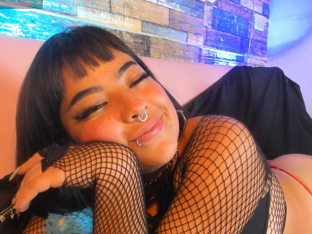 Adarha, con calze a rete nere, piercing al naso e eyeliner, che fa l'occhiolino, è sdraiata, sorride, mostra il seno e indossa una collana nera. La sua pelle è abbronzata ed è carina..