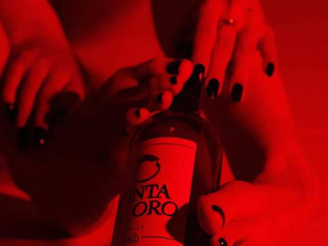 AprillRosen's hands with black nail polish holding a red-lit bottle of Nita Oro, her fingers and ring visible, her bare tits and ass subtly in the background.