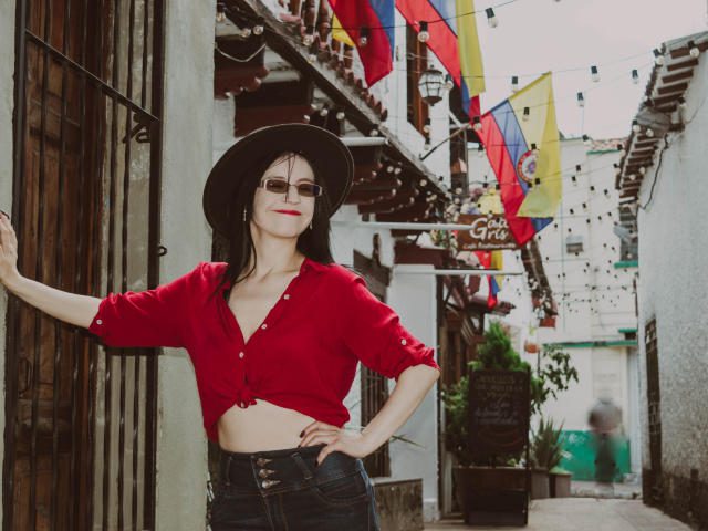 AlanaMoneth with dark hat, red tied-up shirt showing her tits, blue jeans, and sunglasses, standing in a narrow, colorful, Mexican street, arm raised, confident, smiling.