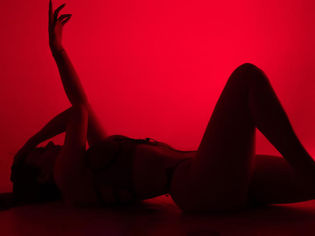 AprillRosen, silhouetteée par une lumière rouge, allongée sur le dos, les bras levés, portant de la lingerie en dentelle noire, mettant en valeur ses fesses et ses seins, dans une pose détendue et séduisante..