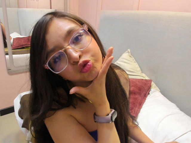 EvaMcAllister, avec des lunettes, faisant la moue avec ses lèvres roses, mimant un baiser, une main levée, de longs cheveux bruns, portant une montre violette, assise sur un lit avec des oreillers blancs..