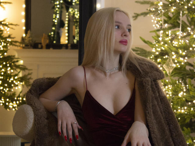 LexyGlam with blonde hair, red lipstick, and red nails, wearing a deep red velvet dress, brown fur coat, and a silver necklace, sitting in a cozy, Christmas-lit room.