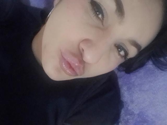 LuluFrancaise has a cute face with shiny pink lips, dark eyebrows, and black hair. She's lying down wearing a black shirt on a purple surface.