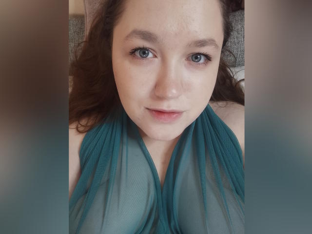 SweetXAliceX mit einem blassgrünen, durchsichtigen Oberteil, das kleine Brüste zeigt, liegend, mit braunen, welligen Haaren, blauen Augen und dezentem Make-up, mit einem sanften Lächeln..