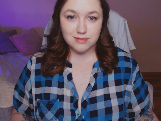 SweetXAliceX has pale skin, blue eyes, and brown wavy hair, wearing a blue and white checkered shirt, looking at the camera with a slight smile.