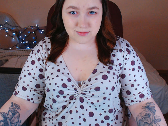 SweetXAliceX mit heller Haut, blauen Augen und braunen, welligen Haaren, trägt ein Punktekleid, zeigt Tattoos an den Armen, sitzt, blickt in die Kamera, leichtes Lächeln..