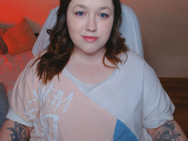 SweetXAliceX tem olhos azuis, pele clara e cabelo castanho ondulado. Ela está vestindo uma camisa branca e rosa com tatuagens nos braços e um leve sorriso..