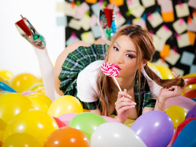 CataleyaLee, avec ses couettes brunes, mange une sucette rose tourbillonnante, allongée dans un tas de ballons colorés, portant une jupe à carreaux verts, une chemise blanche et des talons argentés..
