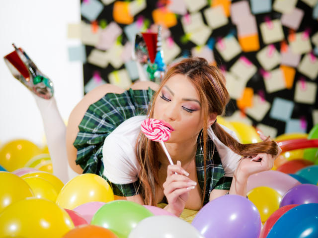 CataleyaLee, avec de grosses seins et une jupe à carreaux, est allongée dans un tas de ballons, en train de sucer un bonbon rose tourbillonnant, et porte des bas blancs montants jusqu'à la cuisse et des talons verts brillants..