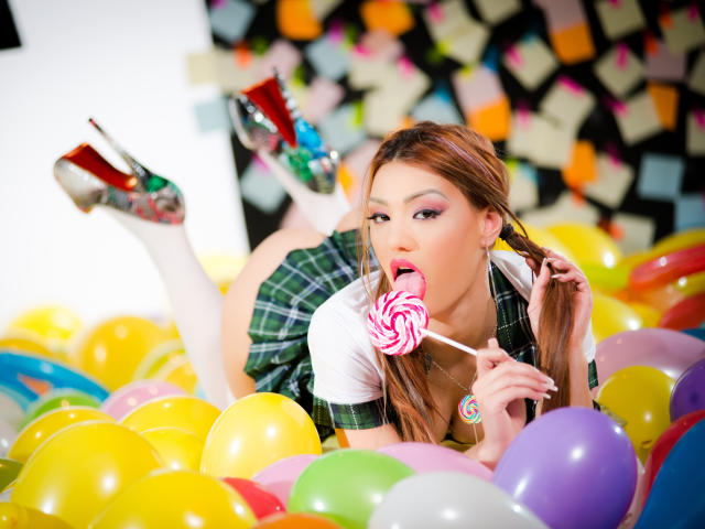 CataleyaLee with brown hair, green plaid skirt, white top, sucking a pink and white swirl lollipop, lying in colorful balloons, wearing white thigh-highs and glittery red heels.