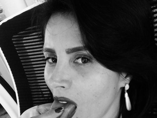 LaurellCharming with dark hair, slightly parted lips, and a pearl earring, biting her finger with black nail polish. Black and white photo.