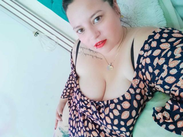 SkayLaraWalker with red lipstick, black patterned dress, off-shoulder, exposing her big tits, tattoo on left shoulder, gold necklace, green pillow, white wall, light skin, dark hair.