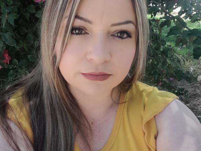 NatalyHami, met lang blond en bruin haar, draagt een gele top, heeft een lichte huid, bruine ogen en donkere lipstick, en kijkt met een lichte glimlach naar de camera..