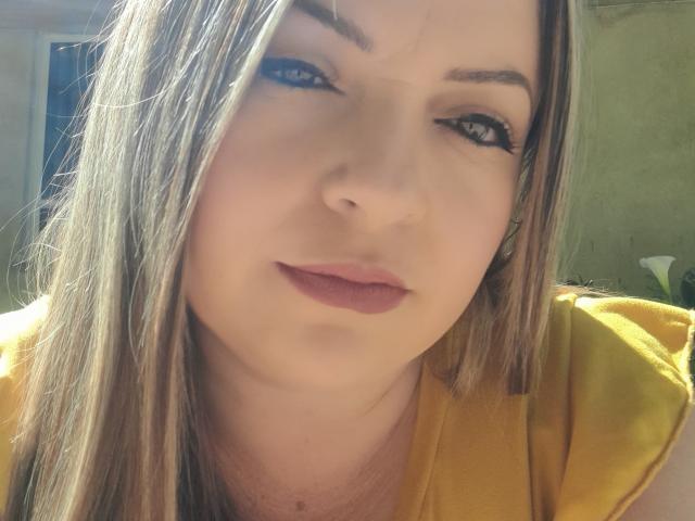 NatalyHami has light brown hair, blue eyeliner, and pink lipstick. She's wearing a yellow shirt, looking slightly to the side with a subtle smile. Her ass is not visible.