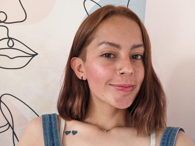Victoria Ortega tiene el cabello castaño claro, un piercing en la nariz y está sacando la lengua con sus labios rosados. Lleva un mono vaquero, mostrando un pequeño tatuaje de corazón en su pecho..