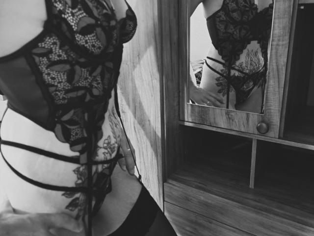 TitaLove en lingerie noire en dentelle, montrant ses seins et ses parties intimes dans un reflet de miroir, avec une porte d'armoire en bois en arrière-plan, portant des bas hauts. Noir et blanc..