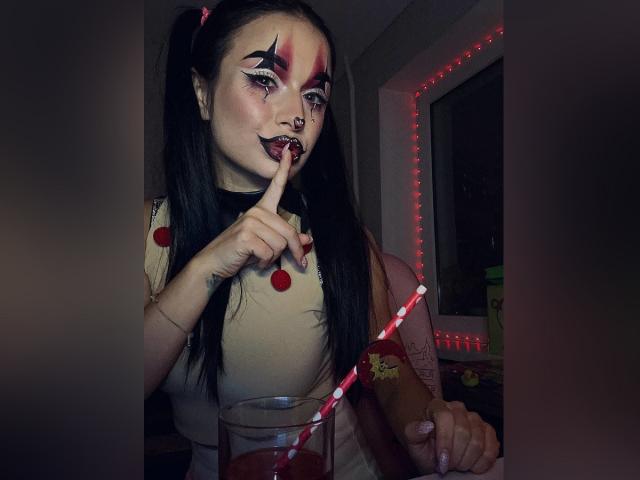 MaryLoveX with dark pigtails, heavy makeup, and red lipstick, shushing with finger to lips, holding a red-striped straw in a glass of red drink, wearing a white top.