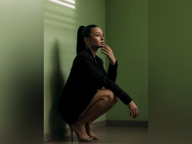 MaryLoveX, squatting, black dress, high heels, hand near mouth, green walls, shadow, looking right, dark brown skin, black ponytail, gold earrings, serious expression, dim lighting.