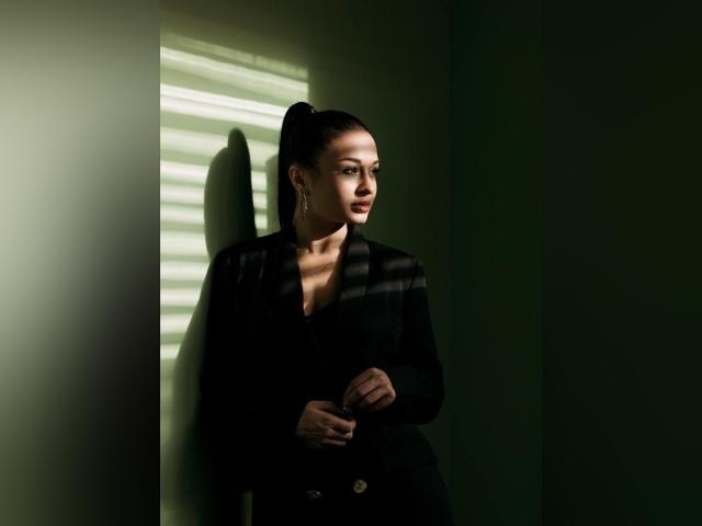 MaryLoveX with dark ponytail, black blazer, and buttons, standing in shadowed green room, light stripes on wall, looking to the side, hands near chest, serious expression.
