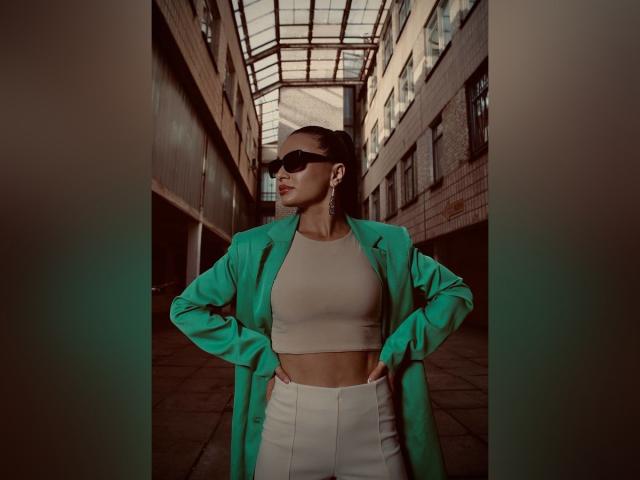 MaryLoveX se tient debout, les mains sur les hanches, portant une veste turquoise par-dessus un crop top beige et un pantalon blanc taille haute, des lunettes de soleil noires et des boucles d'oreilles, dans une ruelle urbaine baignée de soleil..