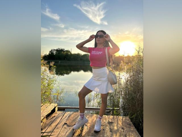MaryLoveX met borsten, gekleed in een roze top, een witte rok en witte sneakers, staat op een houten steiger, met haar handen op haar hoofd, en er is een zonsondergangreflectie in het water te zien. Ze houdt een witte tas vast..