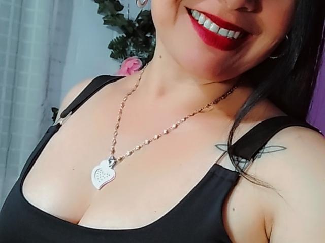 Zaria69 with red lipstick, black tank top, showing her tits, wearing a heart necklace, has a tattoo on her right shoulder, smiling, her face partially visible.