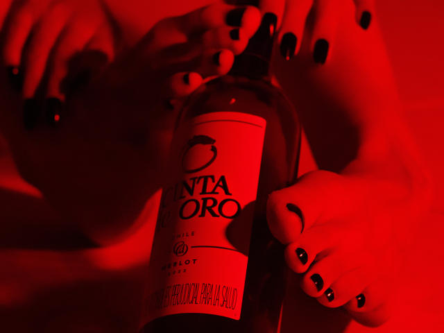 AprillRosen's feet with black nail polish, holding a red-lit bottle of Santa Oro wine, her toes gripping the neck. Red light, wine bottle, and feet, sexy, intimate.