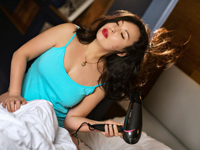 HotLioness with dark hair, red lipstick, and a turquoise tank top, using a black hairdryer on her head while sitting on a bed with white sheets.