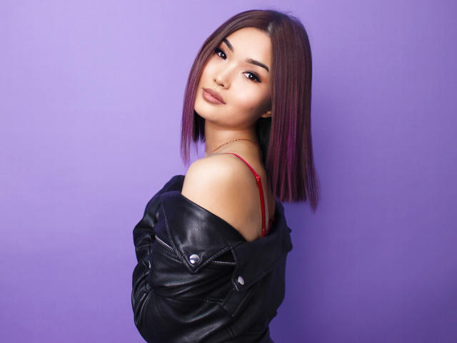 TeyaFaber with purple bob, red bra strap, black leather jacket, slight smirk, purple background, looking over shoulder, soft makeup, subtle eyeliner, gentle smile.