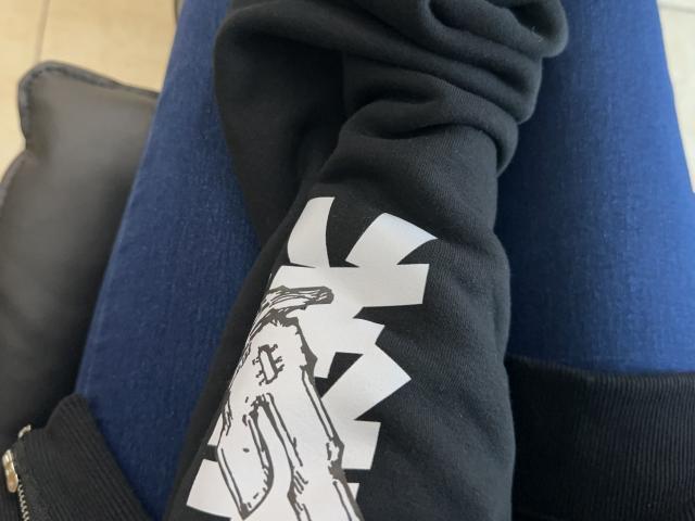 A manga preta da DiLaria com estampa branca, calça jeans azul e bolsa preta. Os seios não estão visíveis. O braço da DiLaria está apoiado em sua perna, que está coberta por jeans..