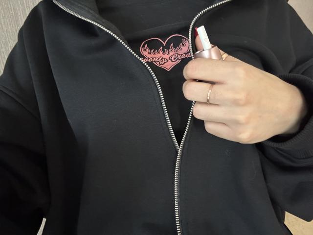 DiLaria's hands zip up a black hoodie, showing a pink heart tattoo with flame details, wearing a silver ring and holding a pink lipstick.