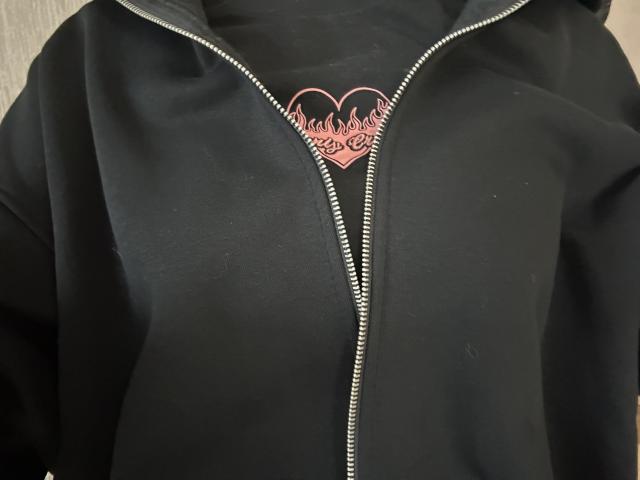 DiLaria's black zip-up hoodie with a shiny metal zipper, featuring a pink heart with flame designs centered on the chest. Boobs not visible, focus on upper torso.